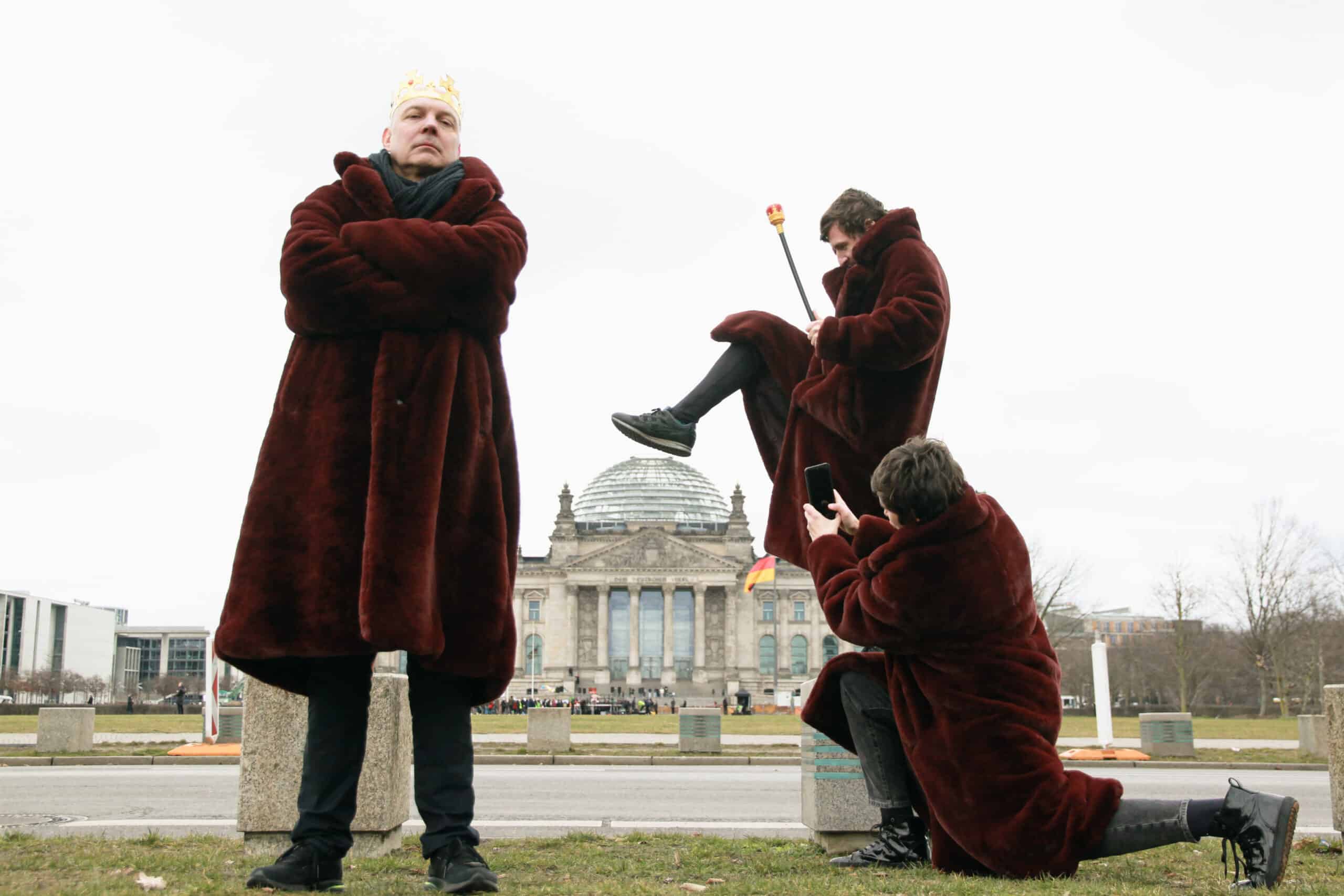3 Personen in pelzähnlichen Mänteln vor dem Reichstagsgebäude. eine Person mit Krone, eione mit Zepter und eine Person macht ein Foto mit dem Smartphone