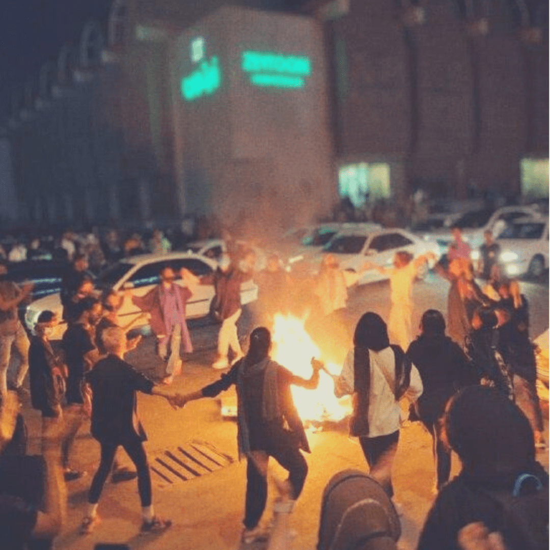 Menschen halten sich an den Händen und tanzen auf dem Bürgersteig im Kreis um ein Feuer. im Hintergrund eine Straße mit vielen Autos und einem großen Gebäude