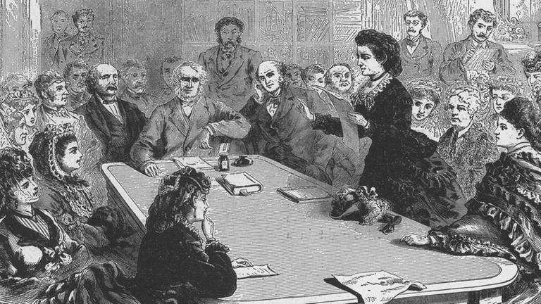 Schwarz-weißes Bild, auf dem eine Frau in alten Gewändern an einem Tisch redet.