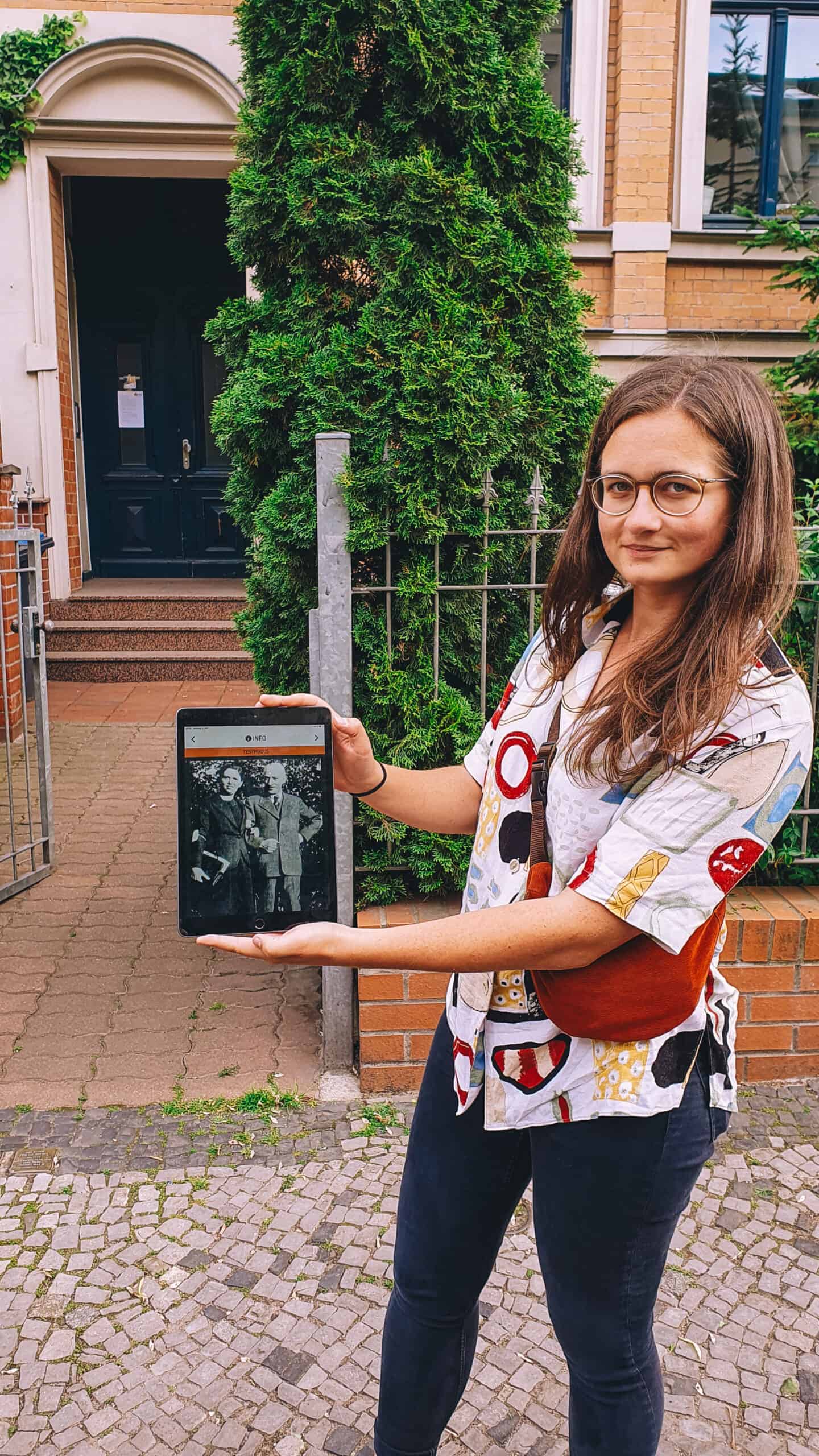 Eine Frau steht vor einem Haus und hält ein Tablet hoch, auf dem ein Bild zu sehen ist