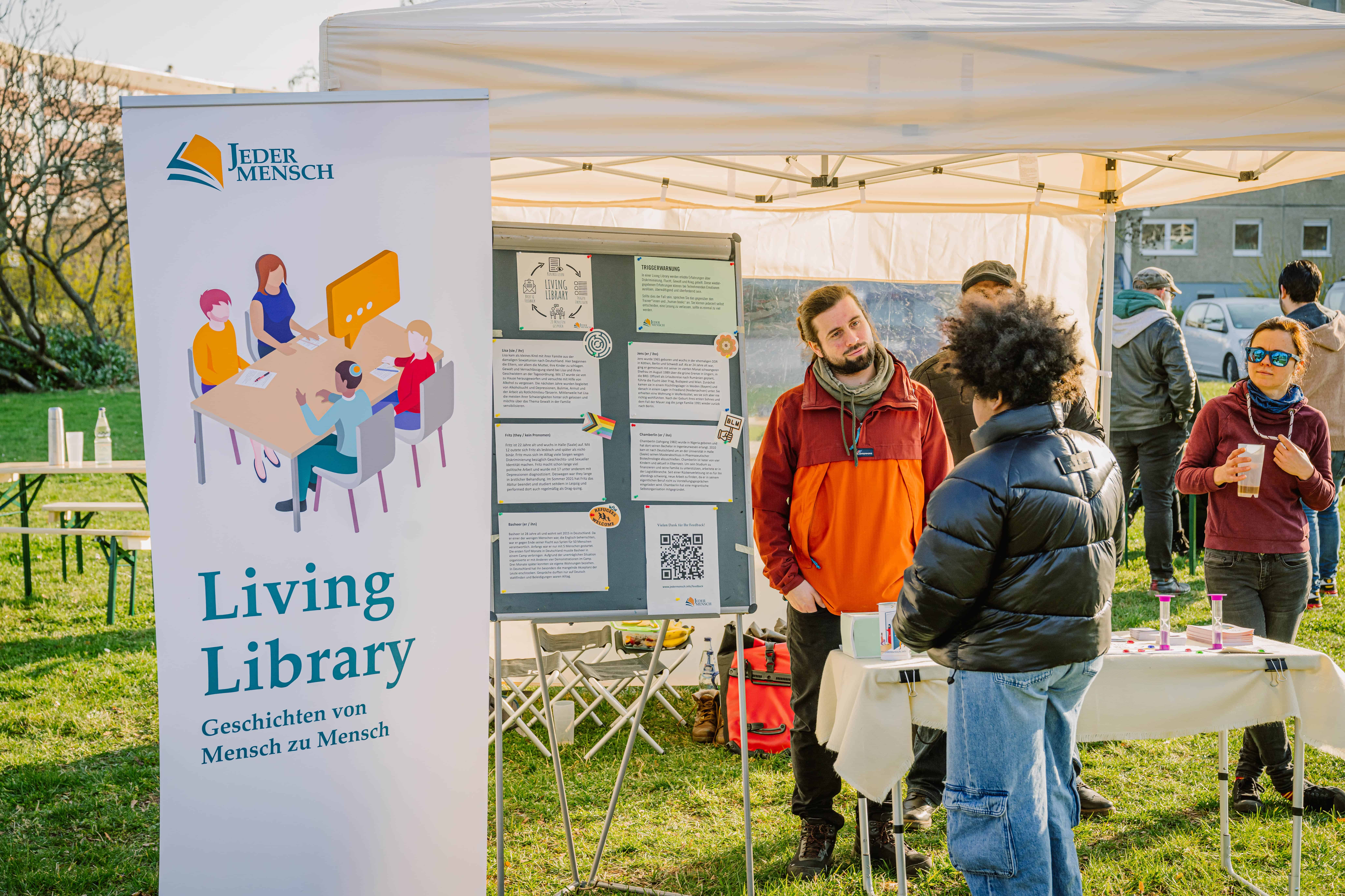 Living Library. Geschichten von Mensch zu Mensch steht auf einem Banner. Zwei Erwachsene unterhalten sich mit einem Kind an einem Info-Stand.